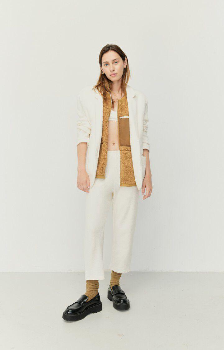 Dame iført hvite joggebukser, beige vest, hvit blazer og sorte sko mot hvit bakgrunn.