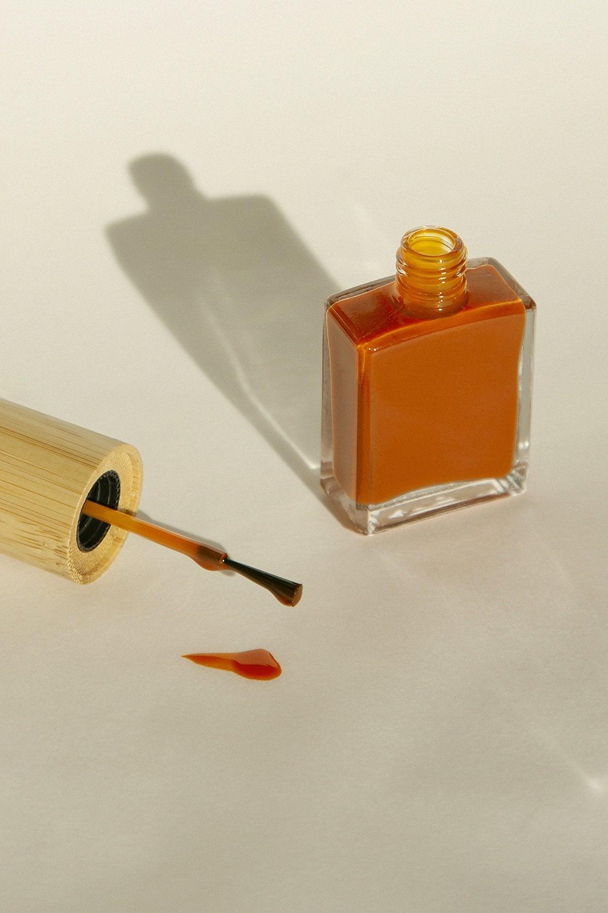 Oransje neglelakkflaske med børste og sølt dråpe på lyst underlag