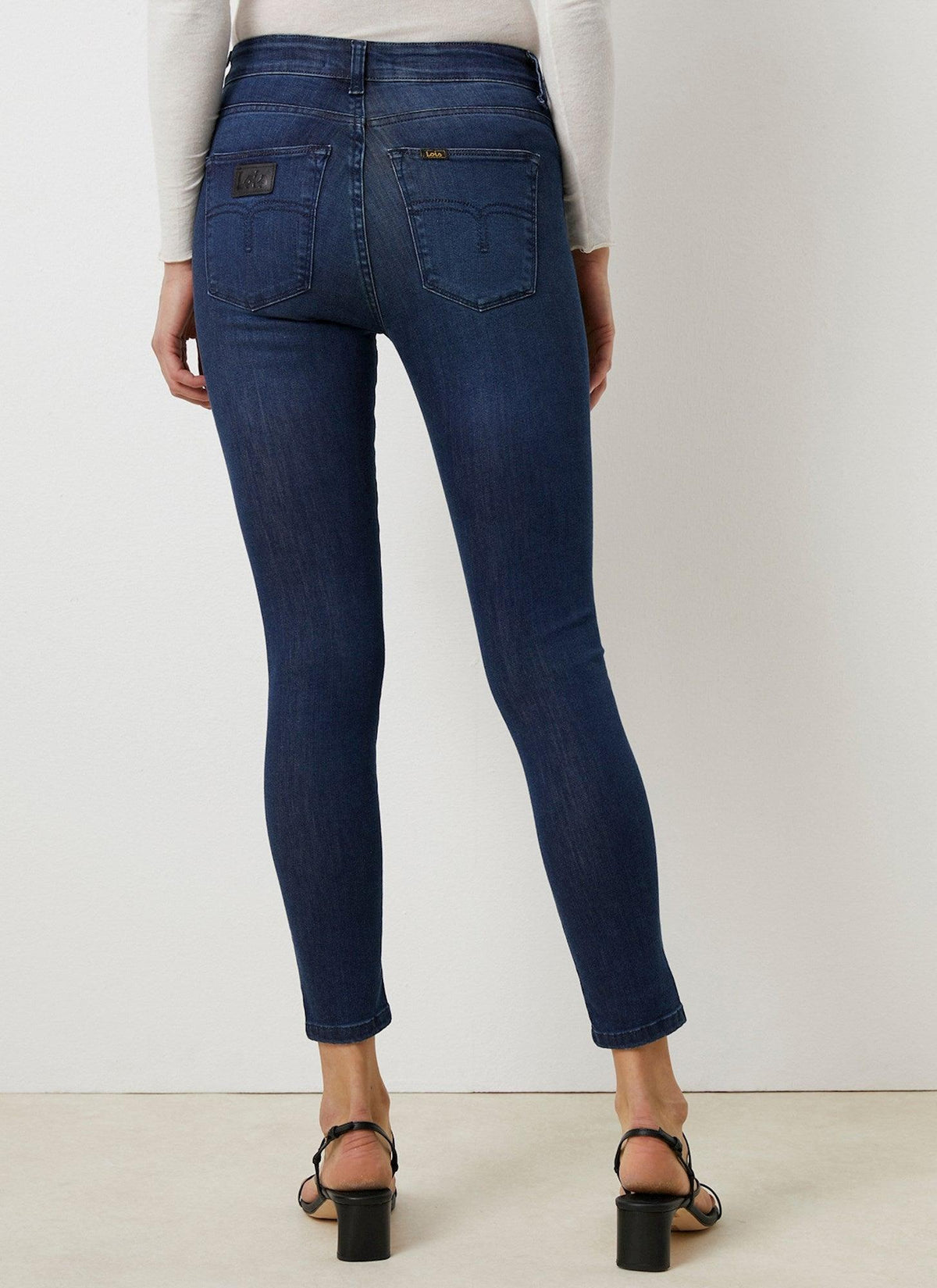 Dame med mørkeblå skinny jeans og sorte sandaler sett bakfra mot hvit bakgrunn