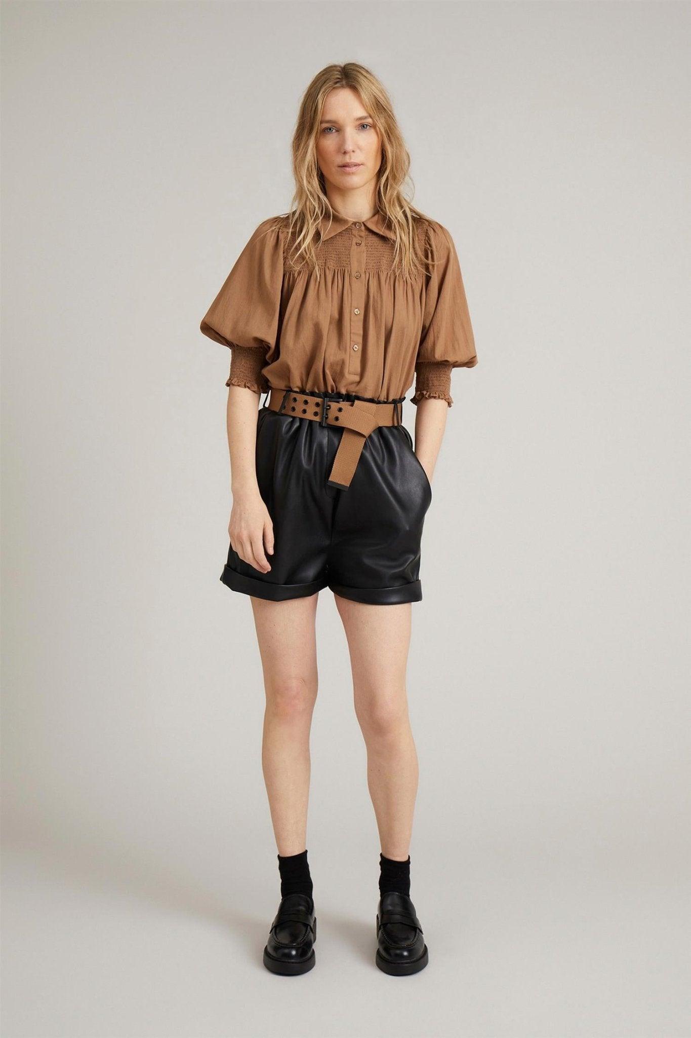 Dame i brune bluse med korte ermer og svarte høytlivs shorts i skinn, belte og loafers
