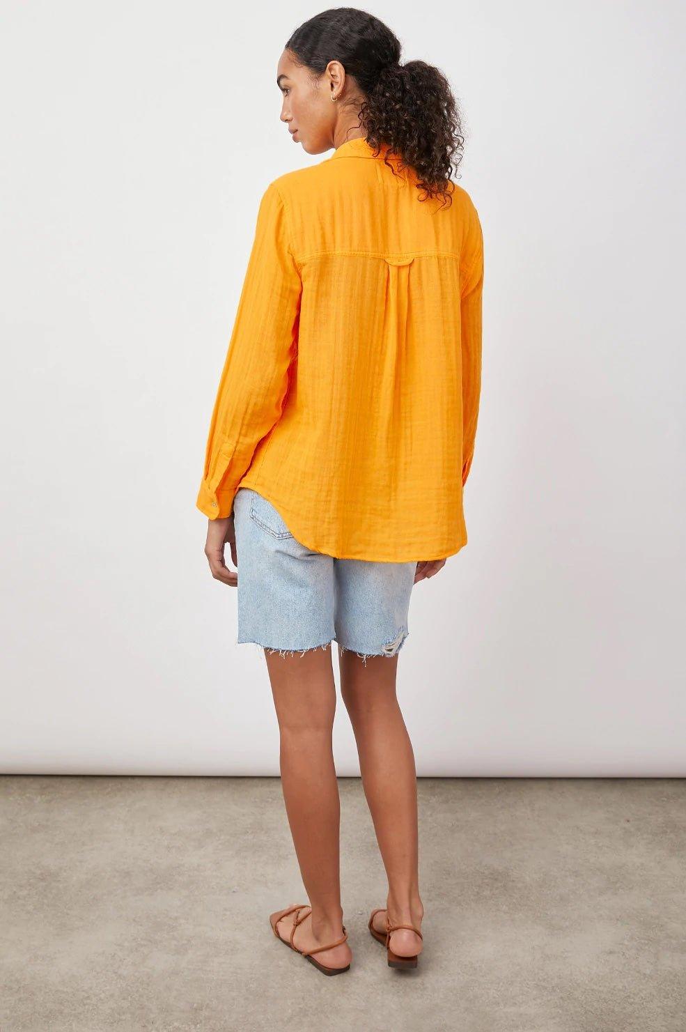 Dame i oransje skjorte og lyse denimshorts, sett bakfra mot hvit bakgrunn.