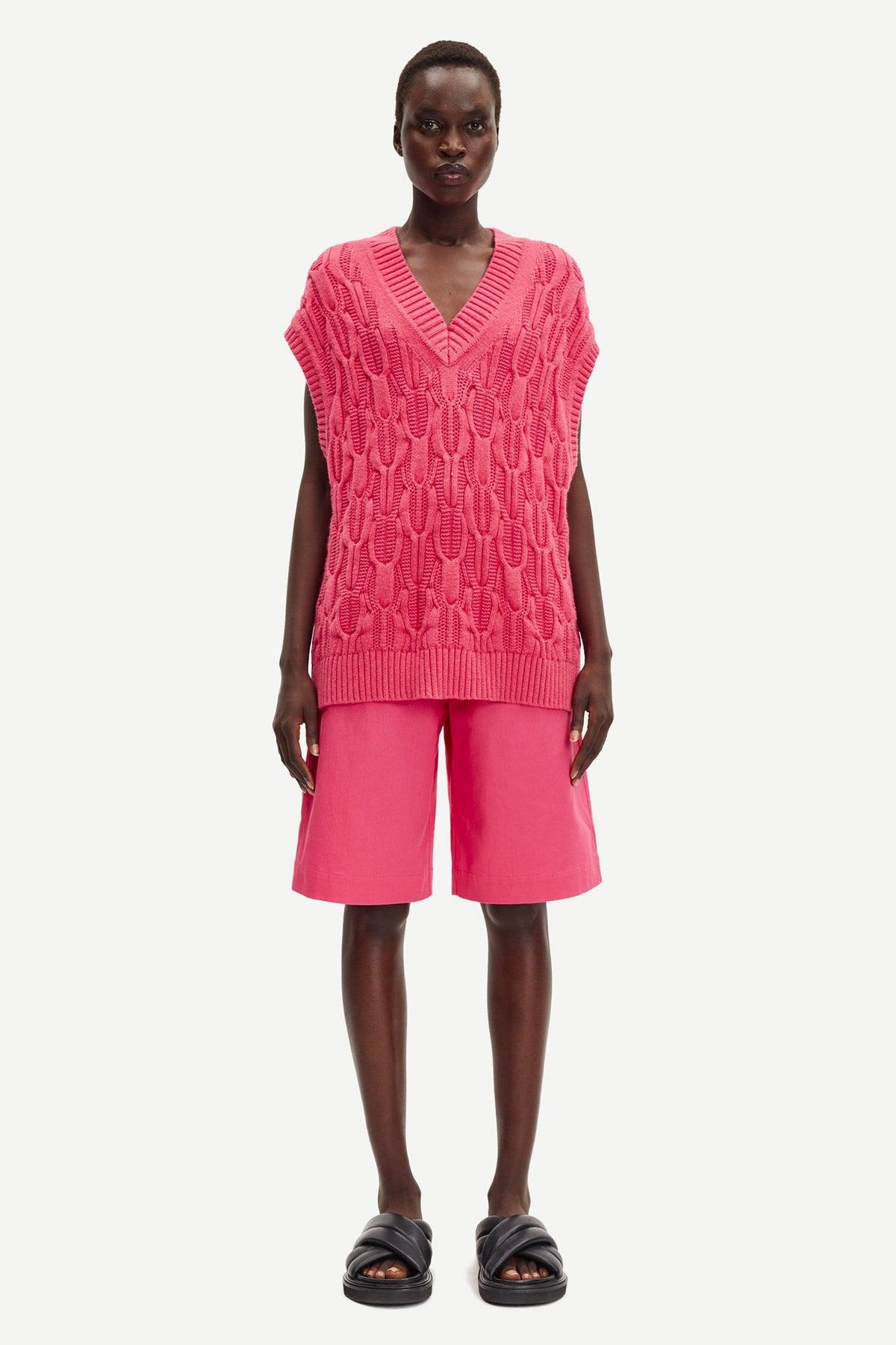 Kvinne iført rosa kabelstrikket vest, rosa bermudashorts og sorte sandaler mot hvit bakgrunn