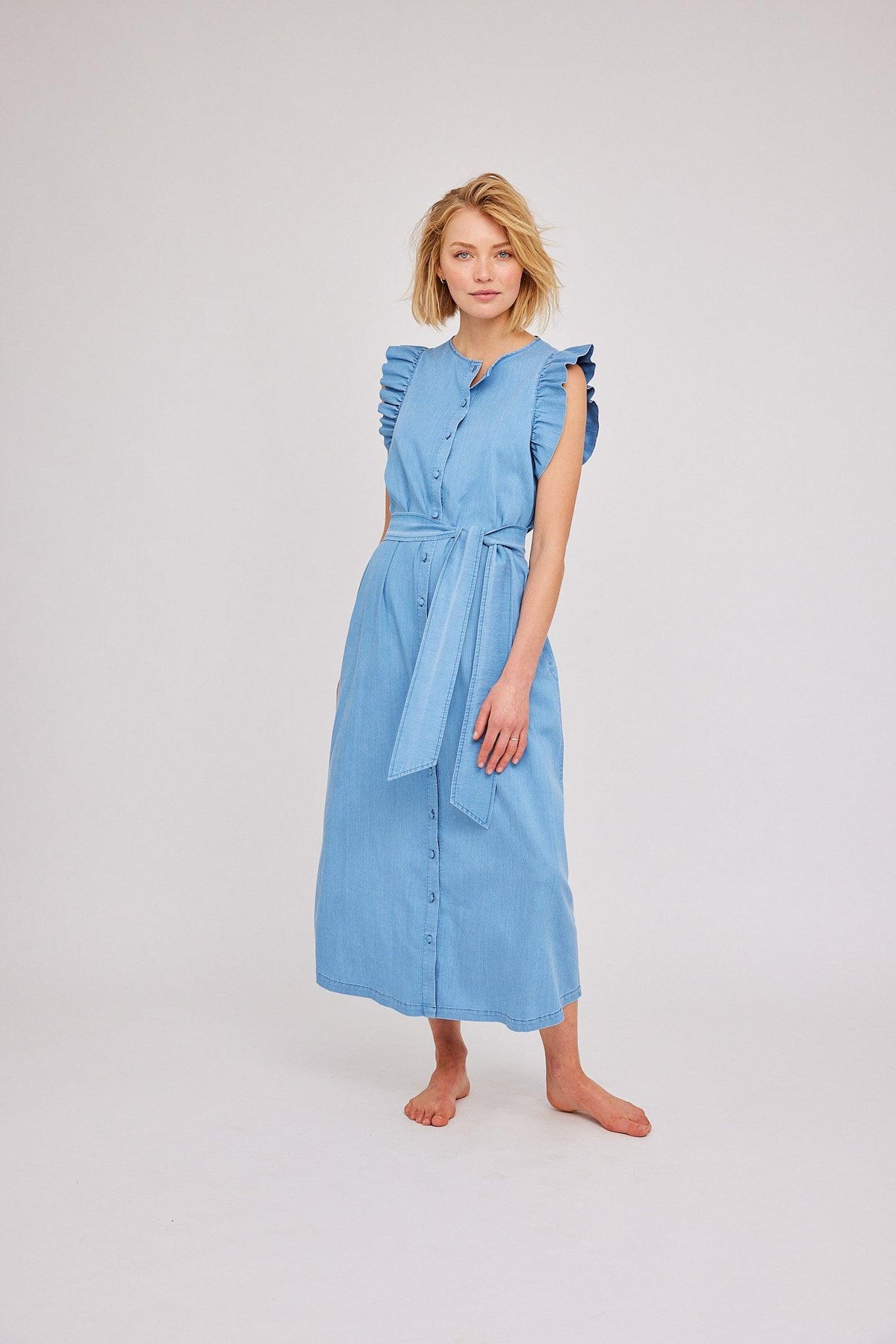 Dame i ermeløs lys blå denim kjole med knapper og belte, mot lys bakgrunn