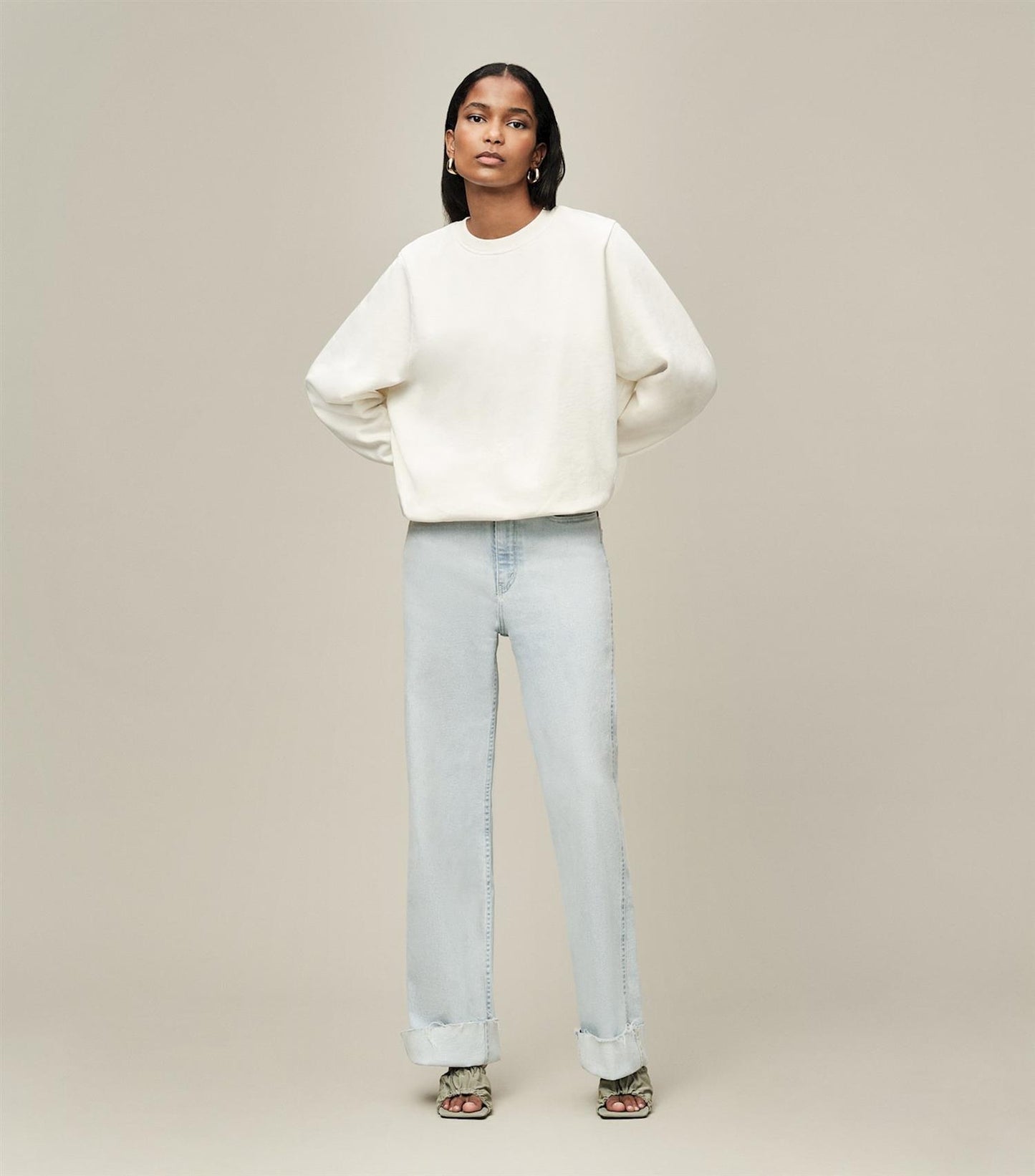 Kvinne i hvit genser og lyseblå jeans med oppbrettede ben, mot nøytral bakgrunn.