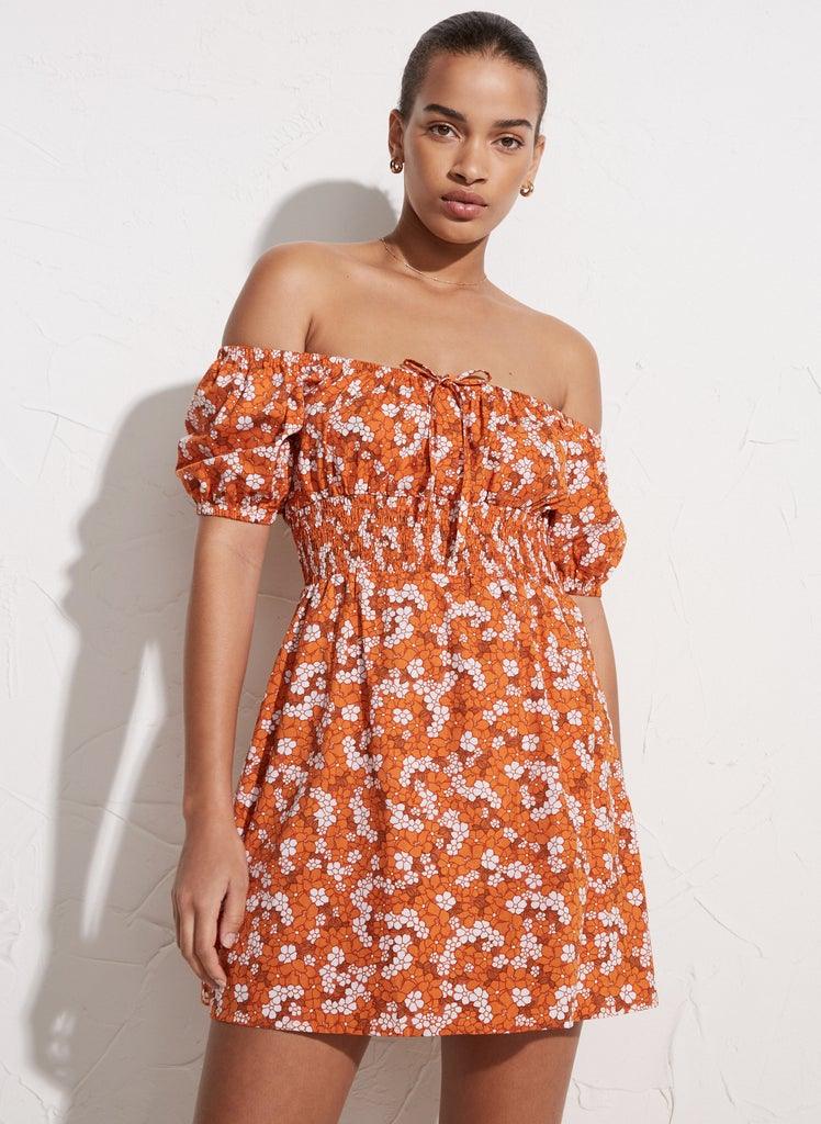 Kvinne med oransje minikjole med hvitt blomstermønster og av-shoulder design mot lys vegg.