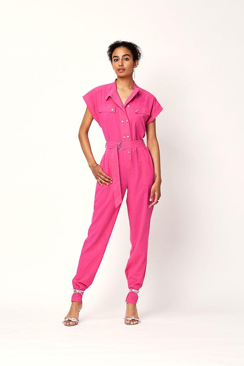 Dame iført magenta jumpsuit med belte og korte ermer, mot lys bakgrunn.