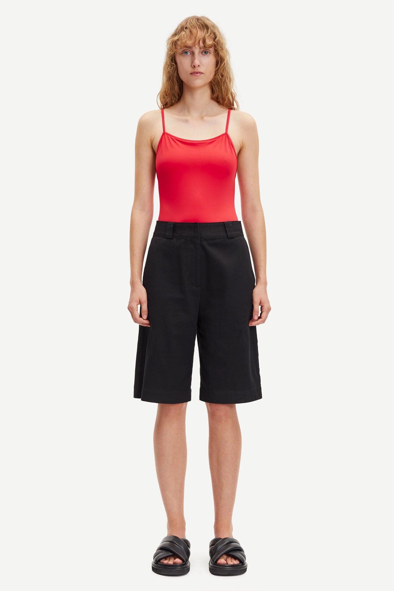Dame i røde singlet og sorte vide bermudashorts, stilrent mot lys bakgrunn