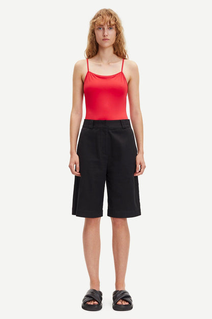 Dame i røde singlet og sorte vide bermudashorts, stilrent mot lys bakgrunn