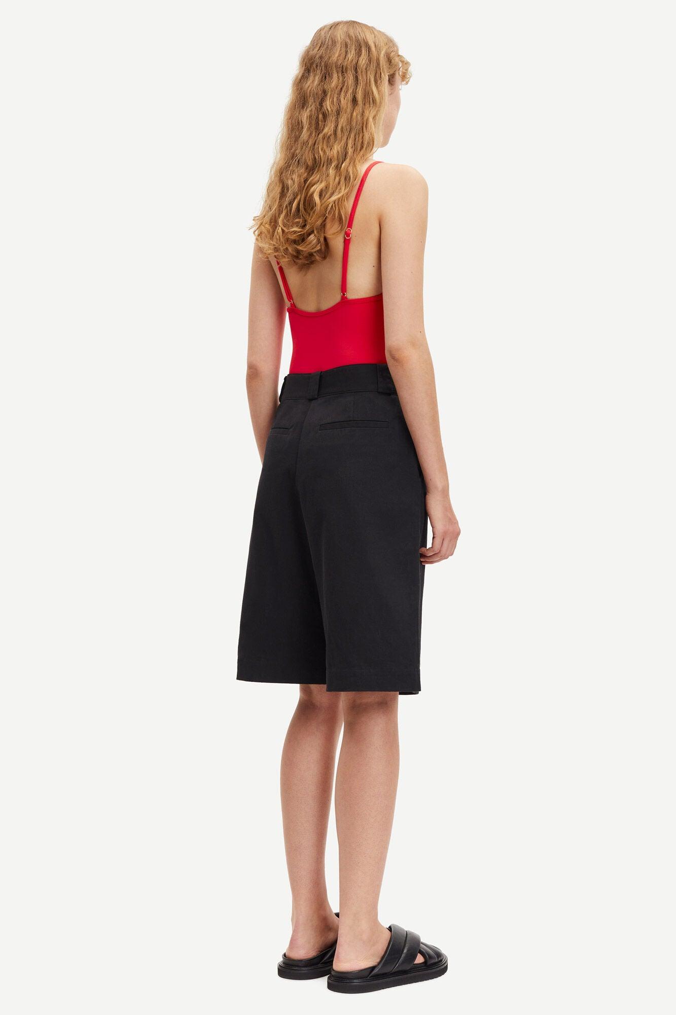 Dame med svarte bermudashorts, rød topp og sorte sandaler, sett bakfra mot hvit bakgrunn.
